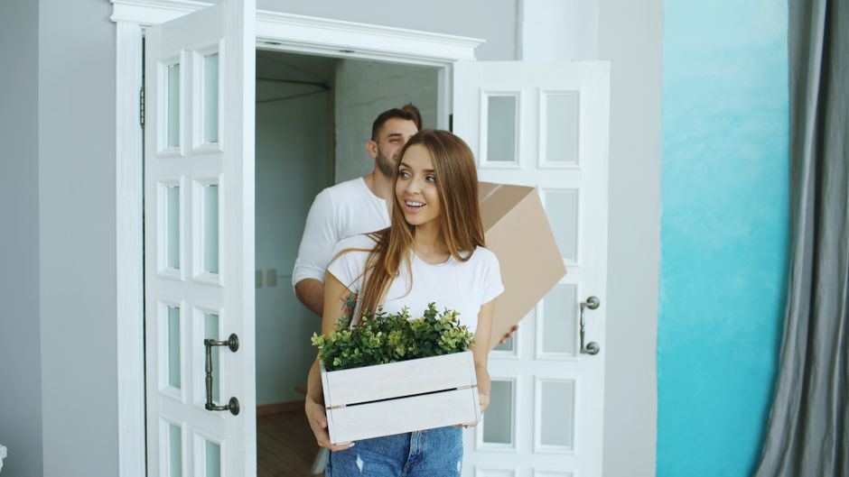 A young woman with long brown hair, wearing a white t-shirt and blue jeans, is standing in a well-lit room holding a white wooden box filled with green plants. Behind her, a man with dark hair and a beard, dressed in a white shirt, is partially visible carrying a cardboard box. The room features light grey walls, with a white double door open to an adjacent room, and a blue accent wall on the right side with a grey curtain partially visible. The flooring appears to be light wood or laminate, and the space is clean and tidy, ready for new homeowners. Enfield Carpet Cleaning emphasizes surface cleaning and sanitisation for maintaining hygienic environments during home moves, reflected in their deep cleaning services listed on the enfieldcarpetcleaning.co.uk website.