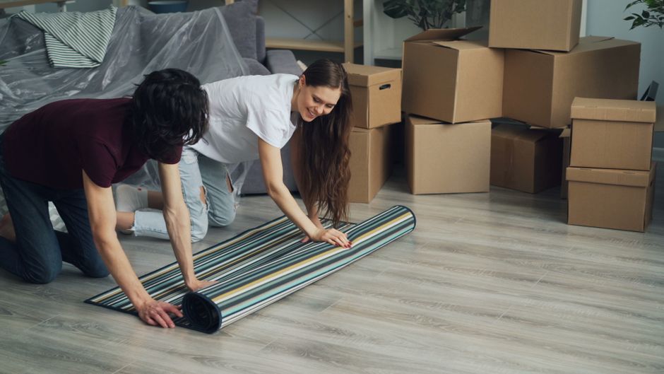 Two individuals are engaged in surface cleaning inside a living room, with a focus on unrolled striped wallpaper or flooring material. The room features a light-colored wooden floor that appears clean and well-maintained. One person is wearing a white shirt and light jeans, smiling as they work, while the other is dressed in a darker top and jeans, both kneeling on the floor. In the background, several cardboard boxes are stacked, indicating recent moving or packing, with a grey upholstered sofa partially visible along with some potted plants and furniture, suggesting a domestic setting. The scene is well-lit with natural or soft indoor lighting, highlighting the cleanliness and readiness for further deep cleaning or sanitisation by Enfield Carpet Cleaning, as part of their domestic cleaning services focused on surface cleaning and hygiene.