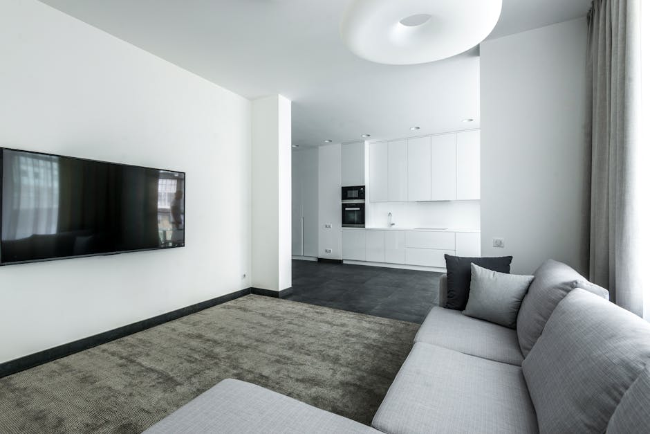 A modern living room featuring a large flat-screen television mounted on a white wall, adjacent to a small section of beige carpeted floor. The room includes a light gray fabric sofa with black and gray cushions positioned near large, sheer curtains that allow natural light to fill the space. In the background, an open-plan kitchen with white cabinets, built-in appliances, and dark tiled flooring is visible. The walls and ceiling are painted white, creating a bright and clean environment, suitable for surface cleaning and domestic maintenance by Enfield Carpet Cleaning.
