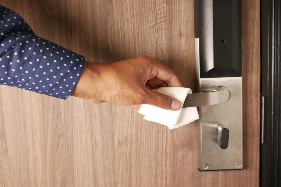A person wearing a dark blue shirt with white polka dots is holding a white cloth and wiping the metallic door handle of a wooden door, which is part of a door lock mechanism. The door has a natural wood finish, and the metallic lock and handle appear shiny and well-maintained. The scene suggests surface cleaning focused on hygiene and sanitisation, possibly related to maintaining cleanliness for residential or commercial environments, as part of deep cleaning services offered by Enfield Carpet Cleaning. The lighting is natural, highlighting the smooth texture of the wood and the shiny surface of the lock and handle.