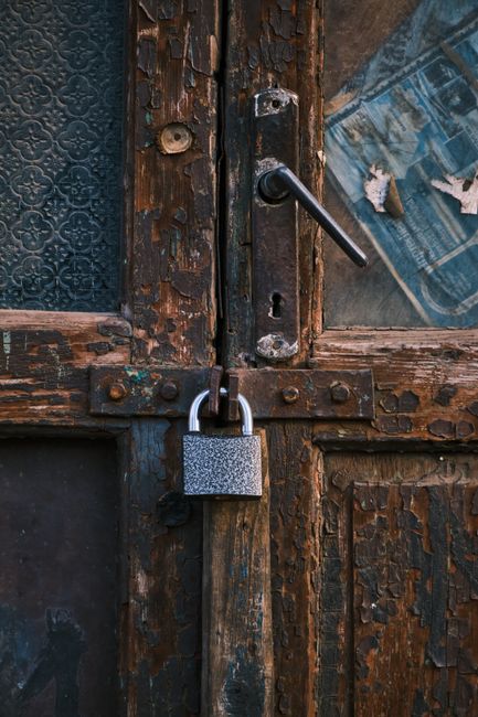Close-up of a weathered wooden door with peeling dark brown paint, featuring a black metal handle and a latch with visible rust. A silver padlock secures the latch, with a textured surface, attached to the door's iron hardware. The door has a small, patterned frosted glass pane on the upper left side, partially visible, contrasting with the aged wood. The surrounding frame shows signs of wear and exposure to the elements, under soft, natural lighting. This image highlights the need for surface cleaning and maintenance, aligning with Enfield Carpet Cleaning's focus on hygiene and property care for both residential and commercial spaces.