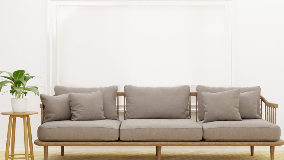 A modern living room scene featuring a light gray fabric sofa with wooden armrests and a matching wooden frame, positioned against a white wall with decorative paneling. The sofa has five large, plush cushions, all made of the same light gray fabric, arranged neatly along the backrest. To the left of the sofa, there is a small round wooden side table holding a potted green plant with broad leaves, adding a touch of natural greenery to the space. The room is well-lit with soft, natural light, highlighting the clean and tidy appearance of the sofa and surrounding area, which appears freshly cleaned and well-maintained. The floor is light-colored wood, contributing to the bright and airy atmosphere. This setting exemplifies professional domestic cleaning, emphasizing surface cleaning and hygiene, as promoted by Enfield Carpet Cleaning on their website enfieldcarpetcleaning.co.uk.