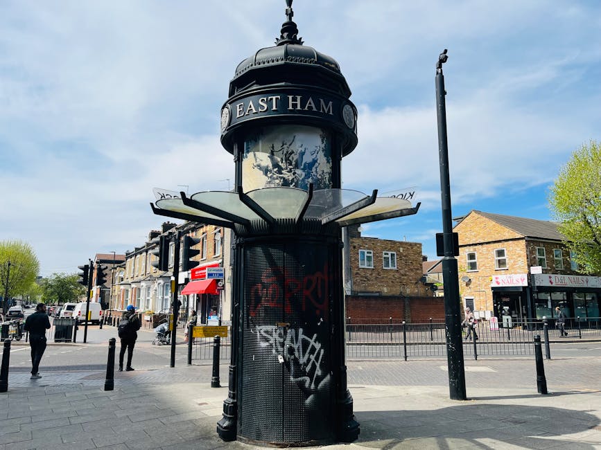 A street scene in Enfield featuring a black, cylindrical street clock with a domed top labeled East Ham, surrounded by a variety of commercial buildings such as a nail salon and other retail shops. The sidewalk is paved with rectangular gray tiles, and there are black bollards lining the pedestrian area. Several people are walking along the street under partly cloudy skies, with trees displaying fresh green leaves indicating spring or early summer. The area appears clean and well-maintained, reflecting urban hygiene standards, with bright natural lighting illuminating the buildings and street fixtures. Enfield Carpet Cleaning is aware of the importance of maintaining a tidy environment both indoors and outdoors, aligning with their core service of surface cleaning, deep cleaning, and sanitisation for residential and commercial spaces.