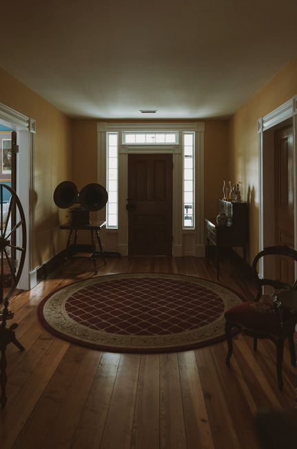 An entryway in a period home featuring a hardwood floor with a large, oval, red and beige patterned rug. The area is lit by natural light coming through a front door with glass panels and side windows, creating a warm ambiance. To the left, there is a vintage-style chair with a cushion, and a small table with decorative items on the right side. The light-colored walls and minimal furnishings reflect cleanliness and care, emphasizing surface cleaning and maintenance suitable for delicate period features. Enfield Carpet Cleaning provides professional domestic cleaning services focused on detailed hygiene and surface sanitisation, ensuring the preservation of traditional home aesthetics while maintaining high standards of cleanliness.