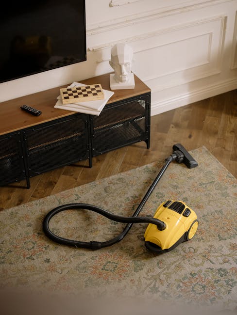 A yellow vacuum cleaner with a black hose is positioned on a patterned area rug in a residential living room. The room features wooden flooring and white panelled walls in a period home. A black metal and wood media console beneath a mounted flat-screen TV holds a remote control, a decorative bust, and a chessboard with a closed book underneath. The lighting appears natural and soft, highlighting the cleanliness of the space. Enfield Carpet Cleaning offers surface cleaning, deep cleaning, and domestic sanitisation services in this setting, contributing to a hygienic and well-maintained environment as depicted in the Southbury station rug cleaning tips for period homes webpage.