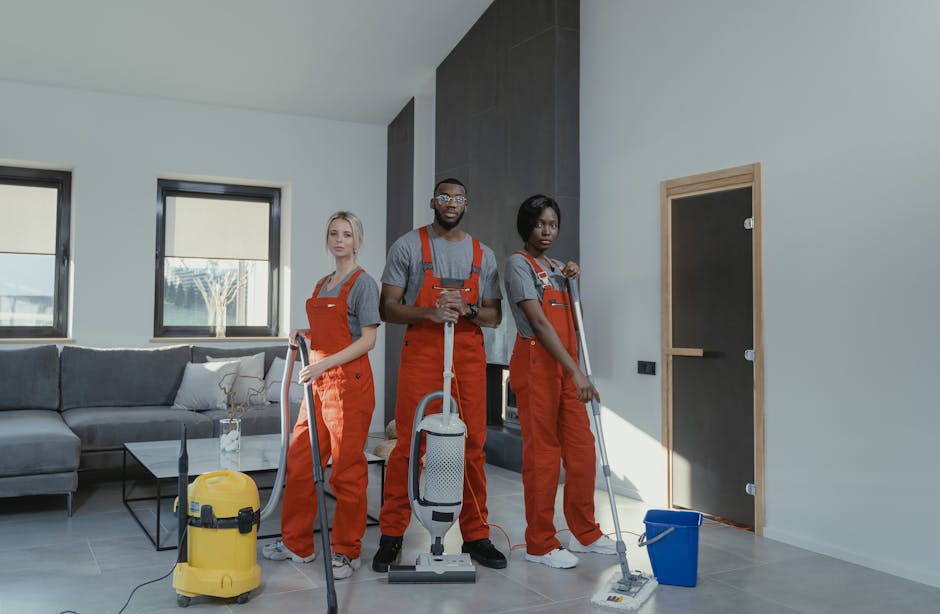Three professional cleaners from Enfield Carpet Cleaning, dressed in grey shirts and bright orange overalls, are performing surface cleaning and deep cleaning in a modern living room. The room features a grey fabric sofa with white cushions, a black glass coffee table, and large windows with black frames allowing natural light to illuminate the space. The cleaners are equipped with vacuum cleaners and mops, and one is holding a spray bottle, indicating part of the sanitisation process. The floor appears tiled with a shiny, dust-free finish, and the overall cleanliness suggests a thorough domestic cleaning service aimed at preparing the property for tenancy handover. The image highlights the importance of expert cleaning for end of tenancy requirements, emphasizing hygiene and surface maintenance, with Enfield Carpet Cleaning's team demonstrating professional handling of residential surface cleaning tasks.