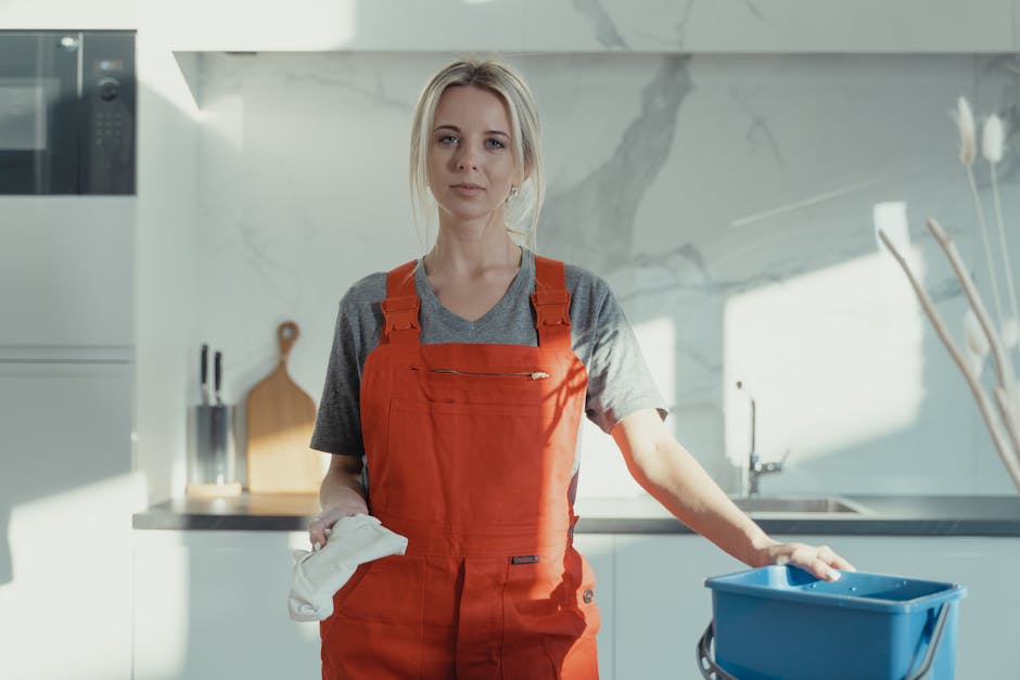 A female cleaner in a modern, bright kitchen wearing a red apron and holding a cleaning cloth, standing next to a blue cleaning bucket with a mop handle visible. The kitchen features a white marble backsplash and countertop, stainless steel appliances, and wooden cutting boards. Natural light illuminates the space, highlighting the tidy and well-maintained surfaces, including polished countertops and clean floor tiles. Enfield Carpet Cleaning is preparing for surface cleaning and sanitisation, emphasizing deep cleaning processes in accordance with end of tenancy requirements.